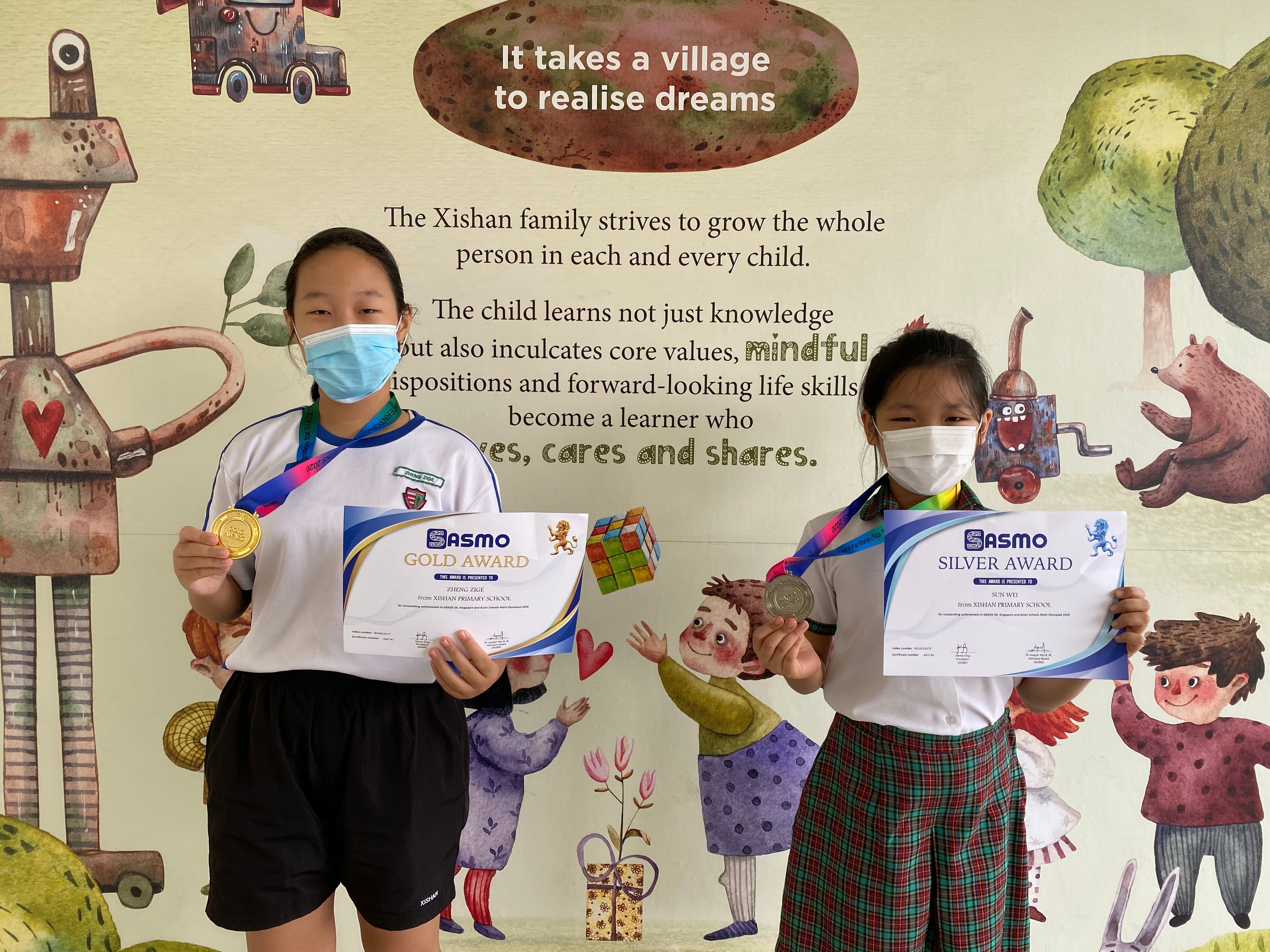 Two masked students hold SASMO Gold and Silver Award certificates with medals in front of a school mural.